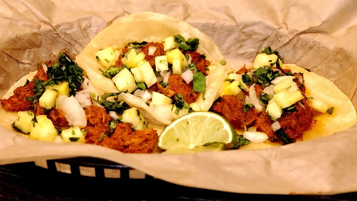 Tacos in a paper-lined basket with chopped toppings and a lime wedge
