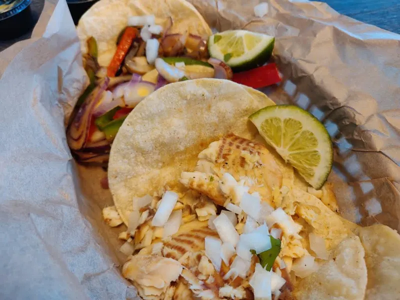 Tacos in a paper-lined basket with grilled peppers and onions, chopped onion, and a lime wedge