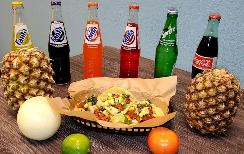Tacos in a basket with bottled sodas, pineapples, an onion, a lime, and a tomato on a wooden table